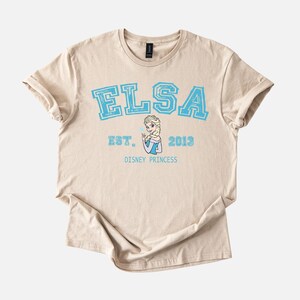 K&ouml;nnte beinhalten: Beiges T-Shirt mit dem Namen "ELSA" in gro&szlig;en, hellblauen Blockbuchstaben. Unter dem Namen steht "EST. 2013" und eine Grafik von Elsa aus Disneys Frozen. Die Worte "DISNEY PRINCESS" sind darunter gedruckt.