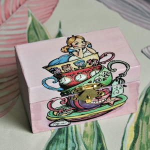 May include: A pink wooden box featuring an Alice in Wonderland design. The box has a gold clasp and is decorated with stacked teacups, Alice, and the Cheshire Cat. The background has a floral pattern.