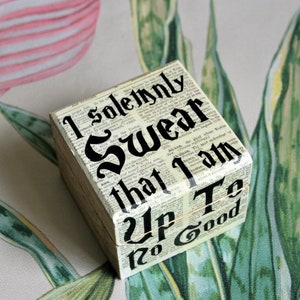 May include: A square wooden box with the text "I solemnly swear that I am up to no good" printed on the sides. The box is decorated with a newspaper print background and black lettering. The box is sitting on a floral patterned surface.