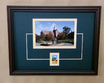 May include: A framed photograph of a building with a clock tower, surrounded by trees and a fountain. The frame is dark brown with a green mat and a white inner mat. A small stamp with a landscape scene is in the lower center of the frame.