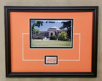 May include: A framed photograph of a building with a white facade and a green lawn in front. The photo is matted with a white mat and a black frame. A small stamp is mounted below the photo.