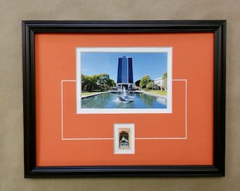 May include: A black framed picture with a matted orange background. The picture is of a building with a fountain in front of it. The building is a modern skyscraper with a reflective glass facade. The fountain is in a reflecting pool. The picture is surrounded by a white mat. There is a smaller picture of a building in the lower right corner of the mat.