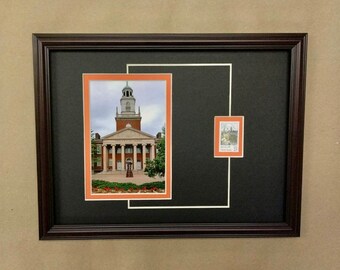 May include: A framed image of a brick building with a clock tower. The building is surrounded by trees and flowers. A smaller image of a stamp with a building is in the upper right corner of the frame.