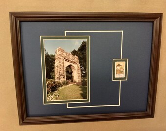 May include: A framed photo of a stone archway with a green lawn and trees in the background. A small postage stamp with a picture of a person is in the lower right corner of the frame.
