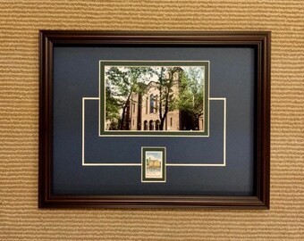 May include: A framed photograph of a building with a dark brown frame and a navy blue mat. The photograph is of a building with a white facade and a green tree in front of it. A small stamp is in the lower right corner of the mat.