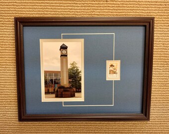 May include: Framed artwork featuring a photograph of a tall clock tower with a clock face, set against a cloudy sky. A postage stamp is also included. The frame is dark brown, with a blue matting.