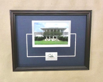May include: Framed photograph of a building with the text "UNIVERSITY CENTER" above the entrance. The photograph is set against a dark blue mat with a white border and a small "LOUISIANA" stamp. The frame is dark brown.