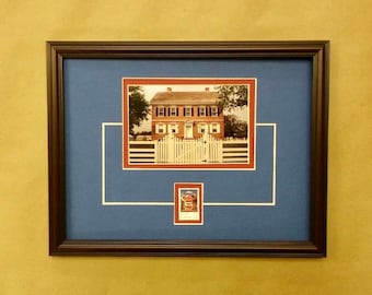 May include: A framed photograph of a brick house with a white picket fence. The house has a red roof and white trim. The photo is matted in blue and has a smaller red and white framed photo of a crest in the lower right corner.