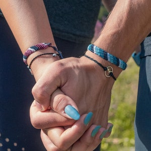 Puede incluir: Dos personas tomadas de la mano, cada una con una pulsera de amistad tejida. Una pulsera es negra, rosa y morada, y la otra es azul y blanca. La persona de la derecha tiene un anillo dorado en su pulsera y esmalte de uñas azul.
