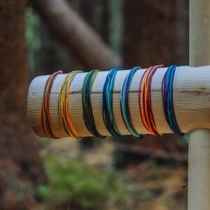 May include: A close-up of a wooden display stand with a variety of colorful bracelets. The bracelets are made of woven string and feature different colors and patterns.