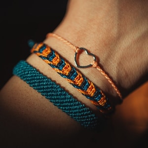 May include: Three bracelets on a wrist, one is a thin orange string with a silver heart charm, one is a thick teal braided cord, and the other is a woven orange and blue bracelet.