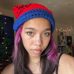 May include: A person wearing a red and blue crocheted beanie with pink highlights in their hair.