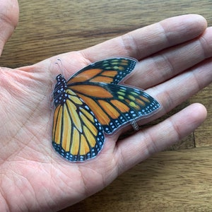 May include: A monarch butterfly sticker with orange, black, yellow, and white details. The sticker is a detailed illustration of the butterfly's wings and body. The sticker is held in a person's hand.