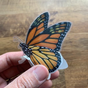 May include: A Monarch butterfly sticker with orange, black, and white details. The butterfly's wings are spread, showcasing intricate patterns and textures. The sticker is held by a hand, with a wooden surface in the background.
