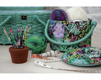 May include: A green and white fabric storage box with a floral pattern of succulents. The box has a handle and is filled with yarn balls. A small brown pot with a green succulent pincushion sits next to the box. A measuring tape and scissors are also visible.