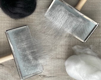 May include: Two rectangular metal hand carders with light-colored wooden handles, featuring fine wire teeth. The carders are positioned with white wool fibers caught between them, alongside a separate ball of black wool and a ball of white wool.