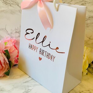 May include: White gift bag with the name "Ellie" and the words "Happy Birthday" in rose gold lettering. A pink ribbon bow adorns the top, and the bag has white rope handles. A small red heart is below the birthday message.