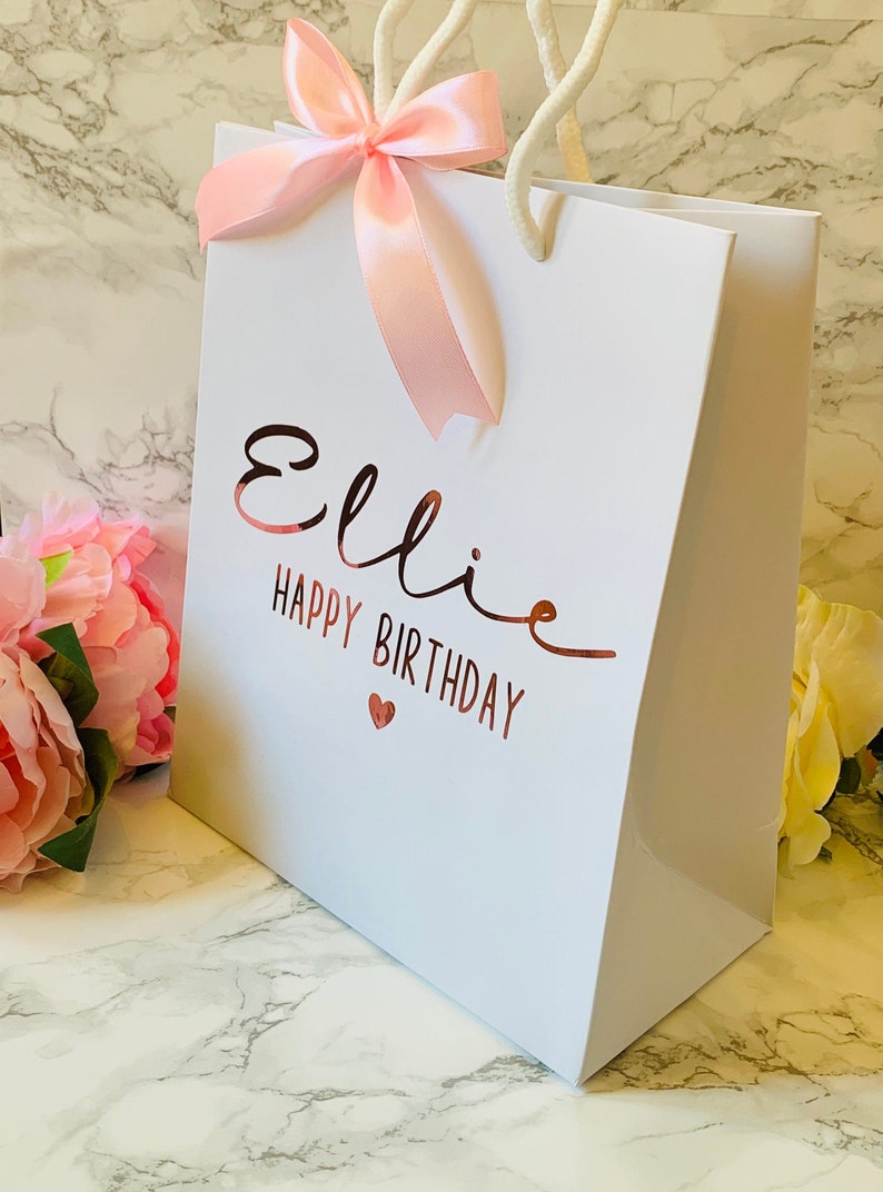 May include: White gift bag with the name "Ellie" and the words "Happy Birthday" in rose gold lettering. A pink ribbon bow adorns the top, and the bag has white rope handles. A small red heart is below the birthday message.