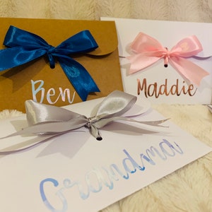 May include: Close-up of three gift envelopes with satin bows. One is brown with a blue bow and the name "Ben". Another is white with a pink bow and the name "Maddie". The third is white with a silver bow and the name "Grandma".