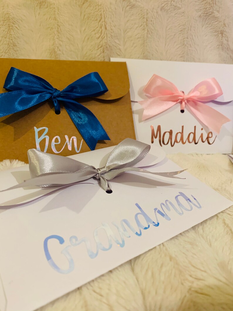 May include: Close-up of three gift envelopes with satin bows. One is brown with a blue bow and the name "Ben". Another is white with a pink bow and the name "Maddie". The third is white with a silver bow and the name "Grandma".