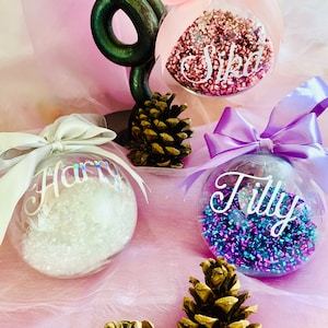 May include: Three clear spherical ornaments filled with colorful shredded material. One contains pink shreds with a pink bow and the name Sika, one has white shreds with a silver bow and the name Harry, and one has blue and purple shreds with a purple bow and the name Tilly.