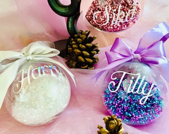 May include: Three clear spherical ornaments filled with colourful shredded material. One contains pink shreds with a pink bow and the name Sika, one has white shreds with a silver bow and the name Harry, and one has blue and purple shreds with a purple bow and the name Tilly.