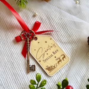 May include: A vintage-style bronze skeleton key tied with a bright red ribbon to a light-colored wooden tag. The tag features the engraved text Santa's SPECIAL KEY, along with small star icons and a graphic illustration of a reindeer-pulled sleigh.