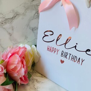 May include: White gift bag with a pink satin bow and rope handles. The bag features the name "Ellie" and the words "Happy Birthday" in a rose gold metallic font, with a small heart. Pink and white flowers are in the foreground.