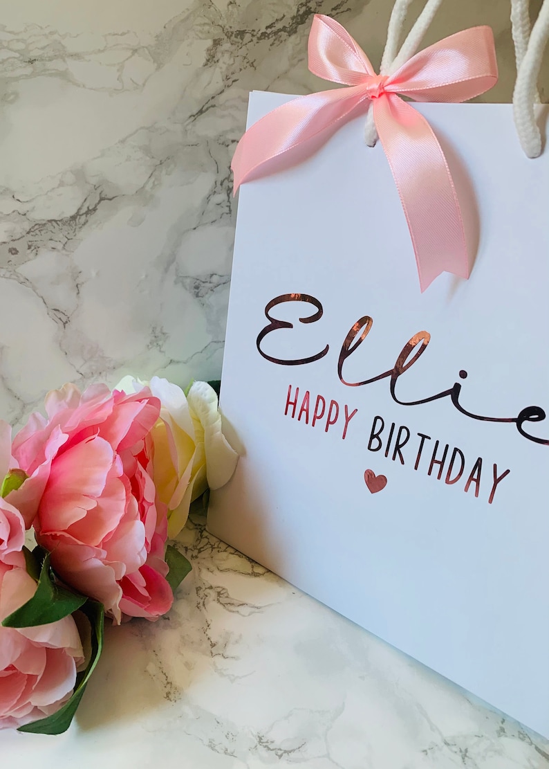 May include: White gift bag with a pink satin bow and rope handles. The bag features the name "Ellie" and the words "Happy Birthday" in a rose gold metallic font, with a small heart. Pink and white flowers are in the foreground.