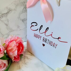 May include: White gift bag with a pink satin bow and white rope handles. The bag has the name "Ellie" and "HAPPY BIRTHDAY" in a metallic rose gold font. Pink peonies and white flowers are in the foreground.