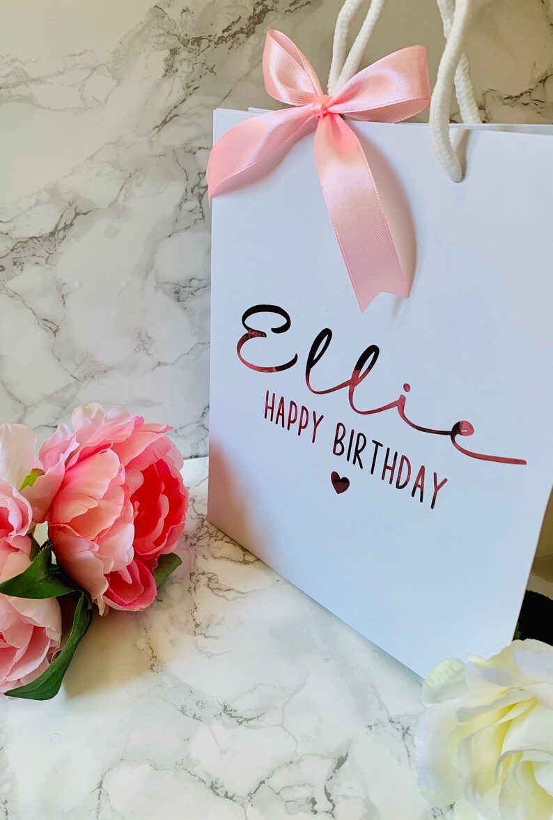 May include: White gift bag with a pink satin bow and white rope handles. The bag has the name "Ellie" and "HAPPY BIRTHDAY" in a metallic rose gold font. Pink peonies and white flowers are in the foreground.