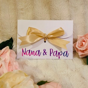 May include: A white envelope with a gold satin bow, featuring the text "Nana & Papa" in a gradient of pink and purple. The envelope is surrounded by pink and cream-colored roses, set against a soft, textured background.