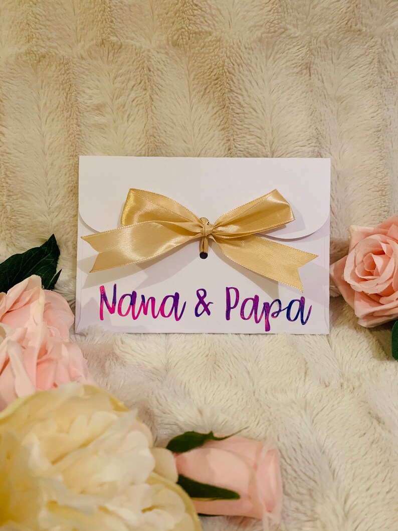 May include: A white envelope with a gold satin bow, featuring the text "Nana & Papa" in a gradient of pink and purple. The envelope is surrounded by pink and cream-colored roses, set against a soft, textured background.
