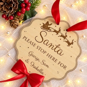 May include: A scalloped wooden sign with a red ribbon hanger and a red bow at the bottom. The sign features a laser-engraved design of Santa in a sleigh with reindeer and the text: Santa PLEASE STOP HERE FOR George, Sam & Isabella.