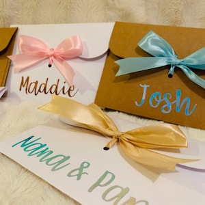 May include: Close-up of personalized gift envelopes with satin bows. The envelopes are white and brown, with names like "Maddie" and "Josh" in metallic script. A tag reads "Nana & Papa". The bows are pink, blue, and gold.