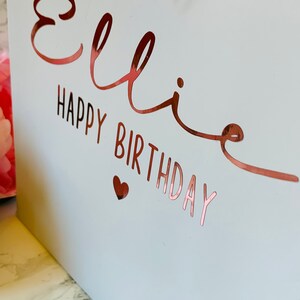 May include: A white gift bag with rose gold lettering that reads "Ellie Happy Birthday" and a small heart. A pink ribbon is tied at the top. The bag is likely for a birthday gift.