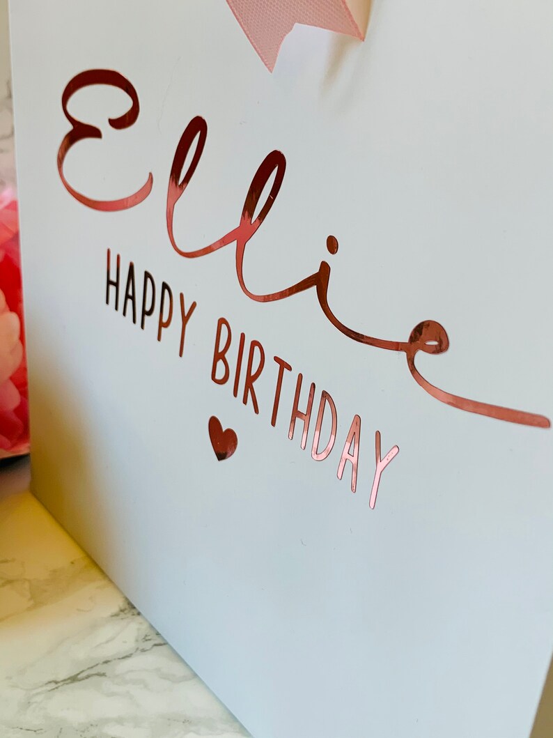 May include: A white gift bag with rose gold lettering that reads "Ellie Happy Birthday" and a small heart. A pink ribbon is tied at the top. The bag is likely for a birthday gift.