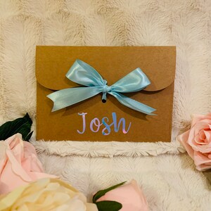 May include: A brown paper gift envelope with a light blue satin bow. The name "Josh" is written on the front in a holographic font. Pink and cream-colored roses are arranged around the envelope.