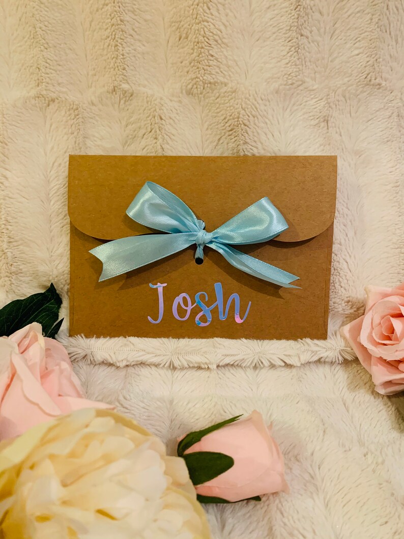 May include: A brown paper gift envelope with a light blue satin bow. The name "Josh" is written on the front in a holographic font. Pink and cream-colored roses are arranged around the envelope.