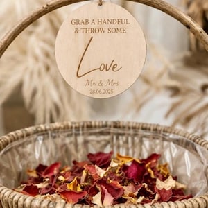 May include: A round light-colored wooden sign hanging from a woven basket handle. The sign features the engraved text: GRAB A HANDFUL & THROW SOME Love Mr. & Mrs. 28.06.2025. The basket is filled with a mixture of dried red, yellow, and white flower petals.