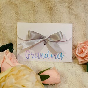 May include: A white card with the word "Grandma" in iridescent script, tied with a silver ribbon bow. The card is surrounded by pink and cream-colored roses, set against a soft, textured background.