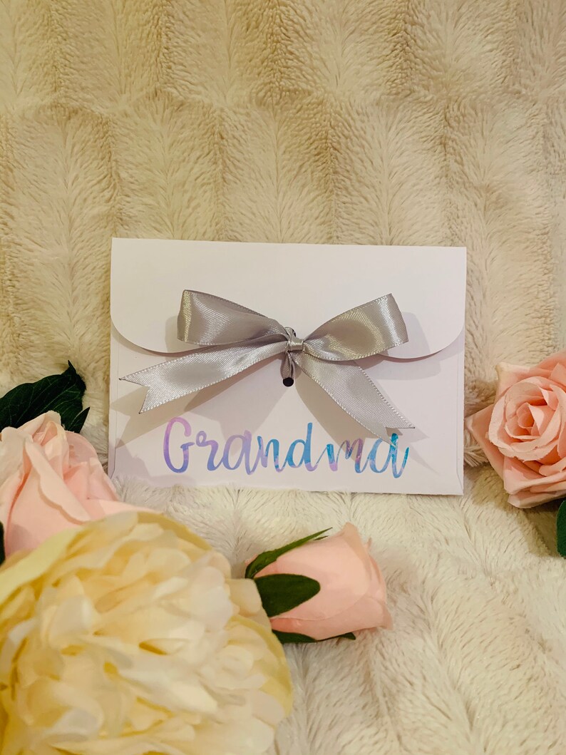 May include: A white card with the word "Grandma" in iridescent script, tied with a silver ribbon bow. The card is surrounded by pink and cream-colored roses, set against a soft, textured background.