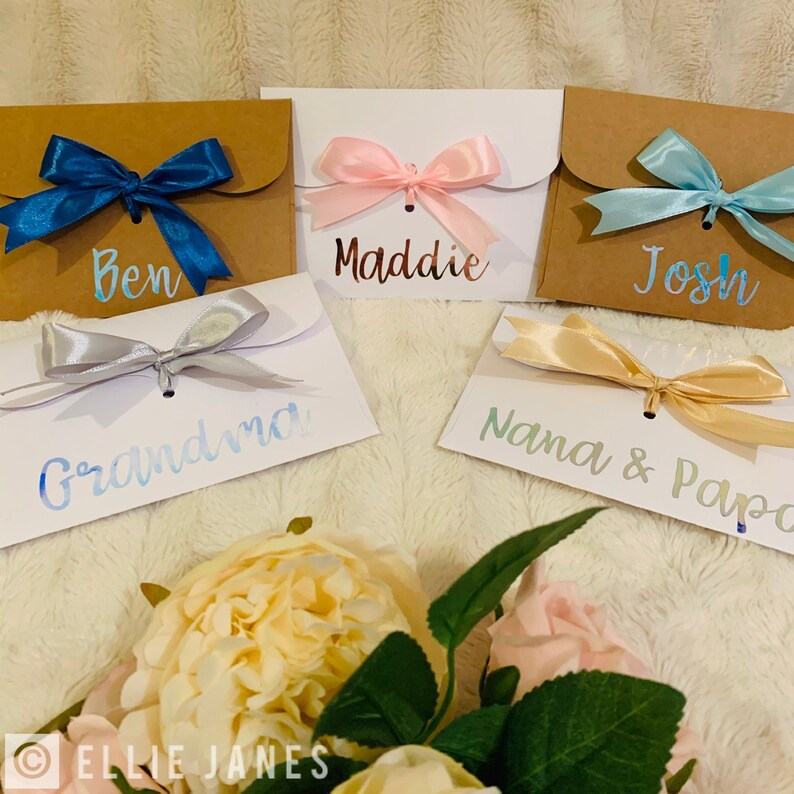 May include: Five small gift envelopes with satin bows. The envelopes are in various colors: brown, white, and kraft. Each envelope has a name written on it: Ben, Maddie, Josh, Grandma, and Nana & Papa. The bows are in shades of blue, pink, silver, and gold.