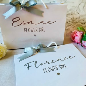 May include: Two white gift bags with gray ribbon bows. One bag reads "Esmae FLOWER GIRL" and the other reads "Florence FLOWER GIRL". Both bags have a small heart graphic. The bags have white rope handles.