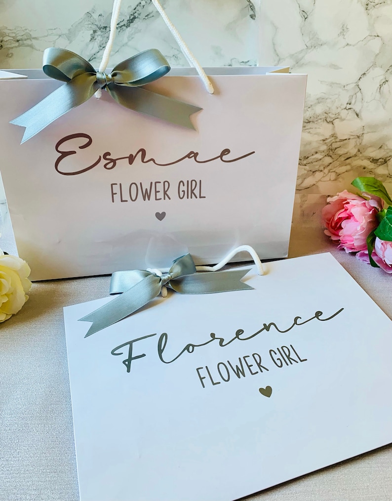May include: Two white gift bags with gray ribbon bows. One bag reads "Esmae FLOWER GIRL" and the other reads "Florence FLOWER GIRL". Both bags have a small heart graphic. The bags have white rope handles.