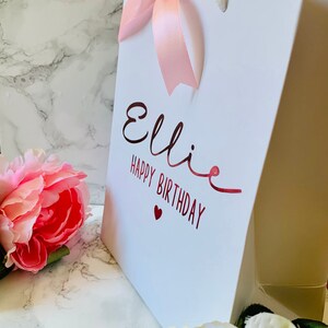 May include: White gift bag with the name "Ellie" and "Happy Birthday" in red script. A pink ribbon bow adorns the top, and a pink flower is in the foreground. The bag has white rope handles.