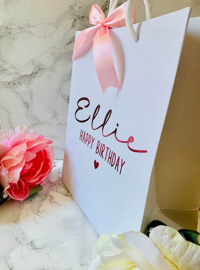 May include: White gift bag with the name "Ellie" and "Happy Birthday" in red script. A pink ribbon bow adorns the top, and a pink flower is in the foreground. The bag has white rope handles.