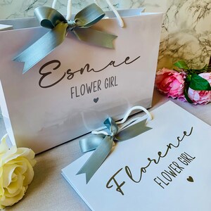 May include: White gift bag and card with the text "Esmae FLOWER GIRL" and "Florence FLOWER GIRL" in elegant script. Each item is adorned with a silver ribbon bow. Pink and yellow flowers are in the background.