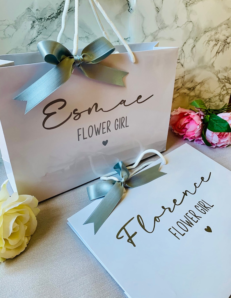 May include: White gift bag and card with the text "Esmae FLOWER GIRL" and "Florence FLOWER GIRL" in elegant script. Each item is adorned with a silver ribbon bow. Pink and yellow flowers are in the background.