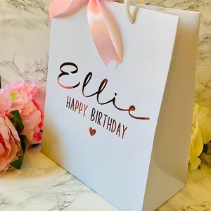 May include: White gift bag with the name "Ellie" and "Happy Birthday" in rose gold script. A pink satin bow adorns the top, with white rope handles. A small red heart is below the birthday message. The bag is on a marble surface.